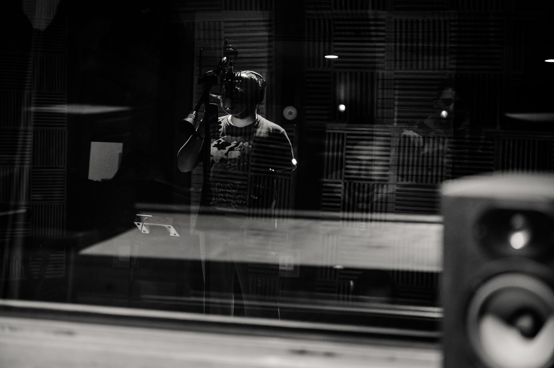 A vocalist at the mic through the booth glass, in black and white